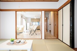 Una habitación tradicional japonesa con suelo de tatami, una mesa baja con juego de té y puertas correderas que dan a una sala de estar con sofá y televisor.
