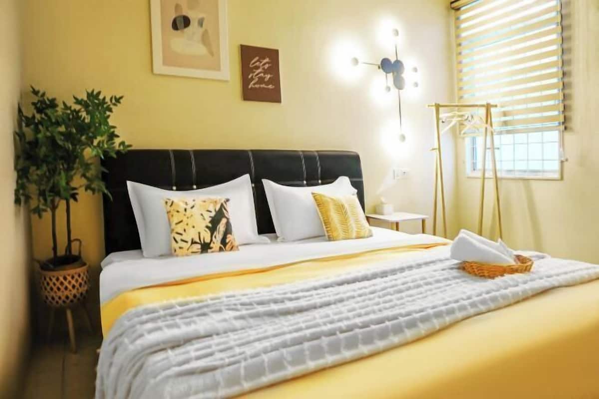 A neatly made bed with a black headboard, a potted plant, and a wall-mounted light fixture.