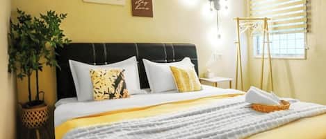 Una cama bien tendida con cabecera negra, una planta en maceta y una lámpara fijada a la pared.