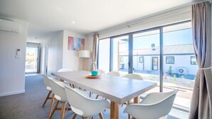A modern dining area with a large table and white chairs, large windows with a view of buildings, and a lamp on the wall.