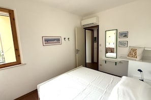 A bedroom with a bed, a window, a door, a mirror, and a wall-mounted shelf with a pillow.