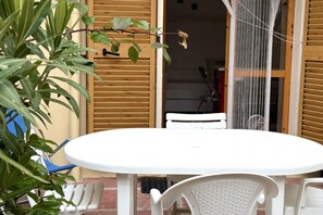 A white table and chairs outside a building with wooden shutters, a hanging plant, and a mesh screen.