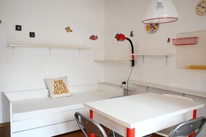 A room with a white table, a white bench with a pillow, a red lamp, and wall decorations including fish and a clock.
