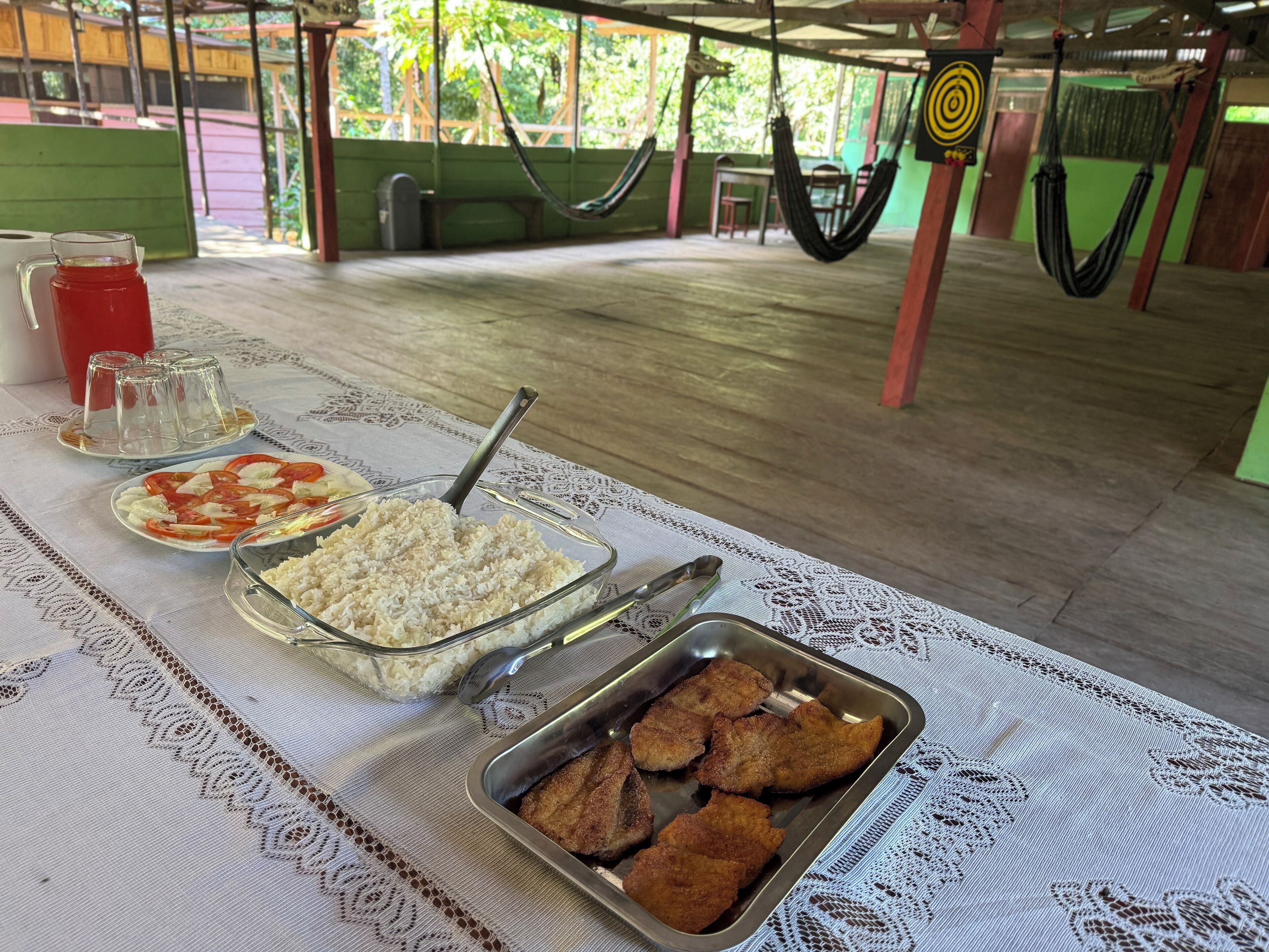 Uma mesa de jantar com comida e bebidas, um redes e uma cadeira de balanço ao fundo.