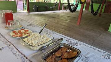 Uma mesa de jantar com comida e bebidas, um redes e uma cadeira de balanço ao fundo.