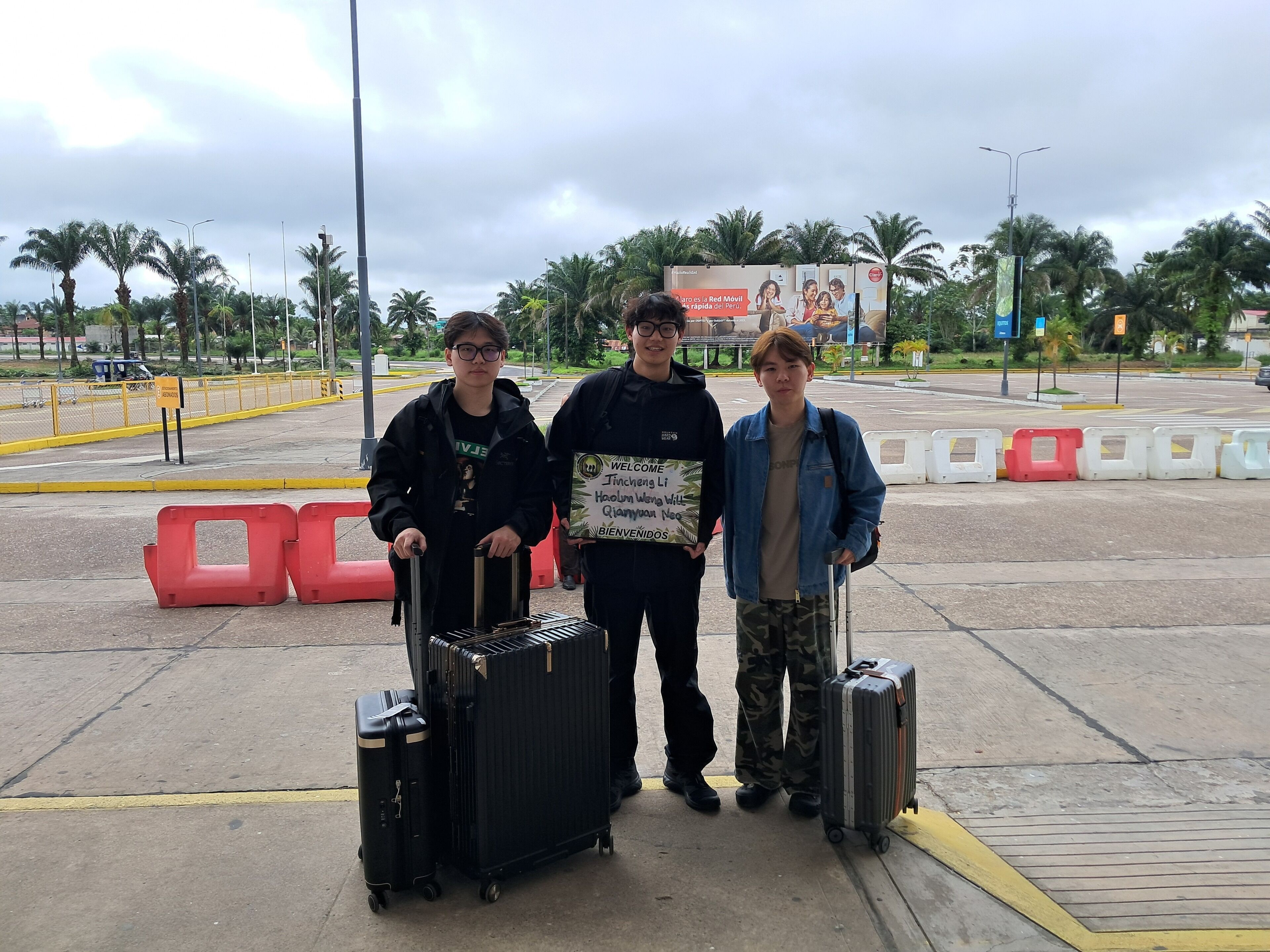 Three individuals standing in an outdoor area with luggage, palm trees, and a signboard in the background.