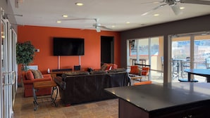 Una sala moderna con un televisor grande, asientos cómodos, un ventilador de techo y vistas amplias a través de ventanas grandes.