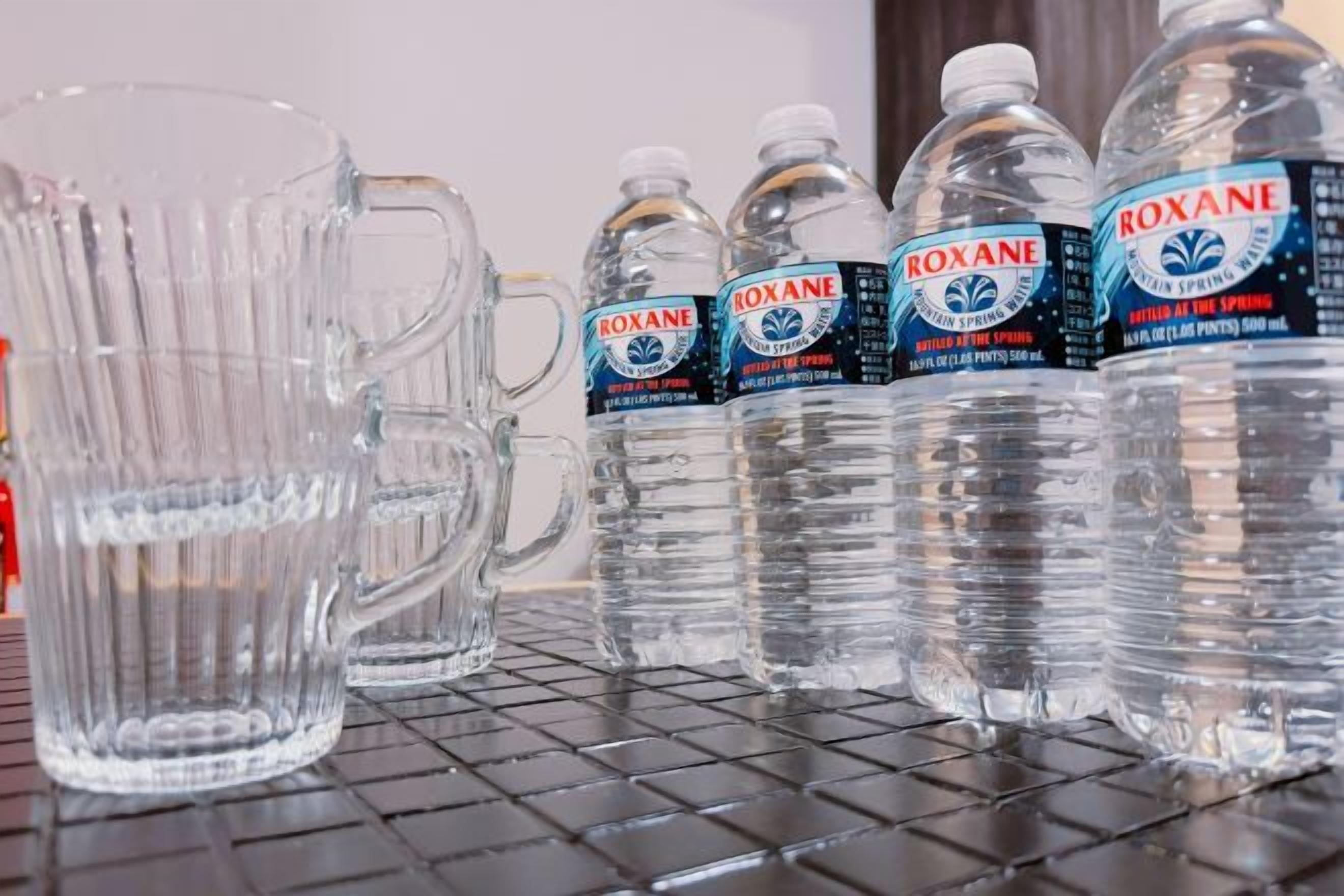 A row of clear glass mugs and four bottles of Roxane water on a shelf.