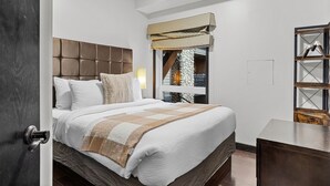 Ein Schlafzimmer mit einem Bett, einem Nachttisch, einem Fenster mit Blick auf eine Steinmauer und ein Holzgebäude im Freien sowie einer Tür links.
