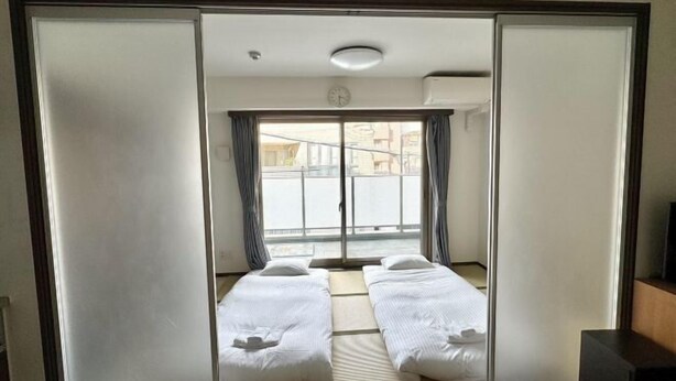 A room with a sliding door, a bed with white bedding, a window with curtains, and a clock on the wall. - Jin Yue guesthouse (Osaka)