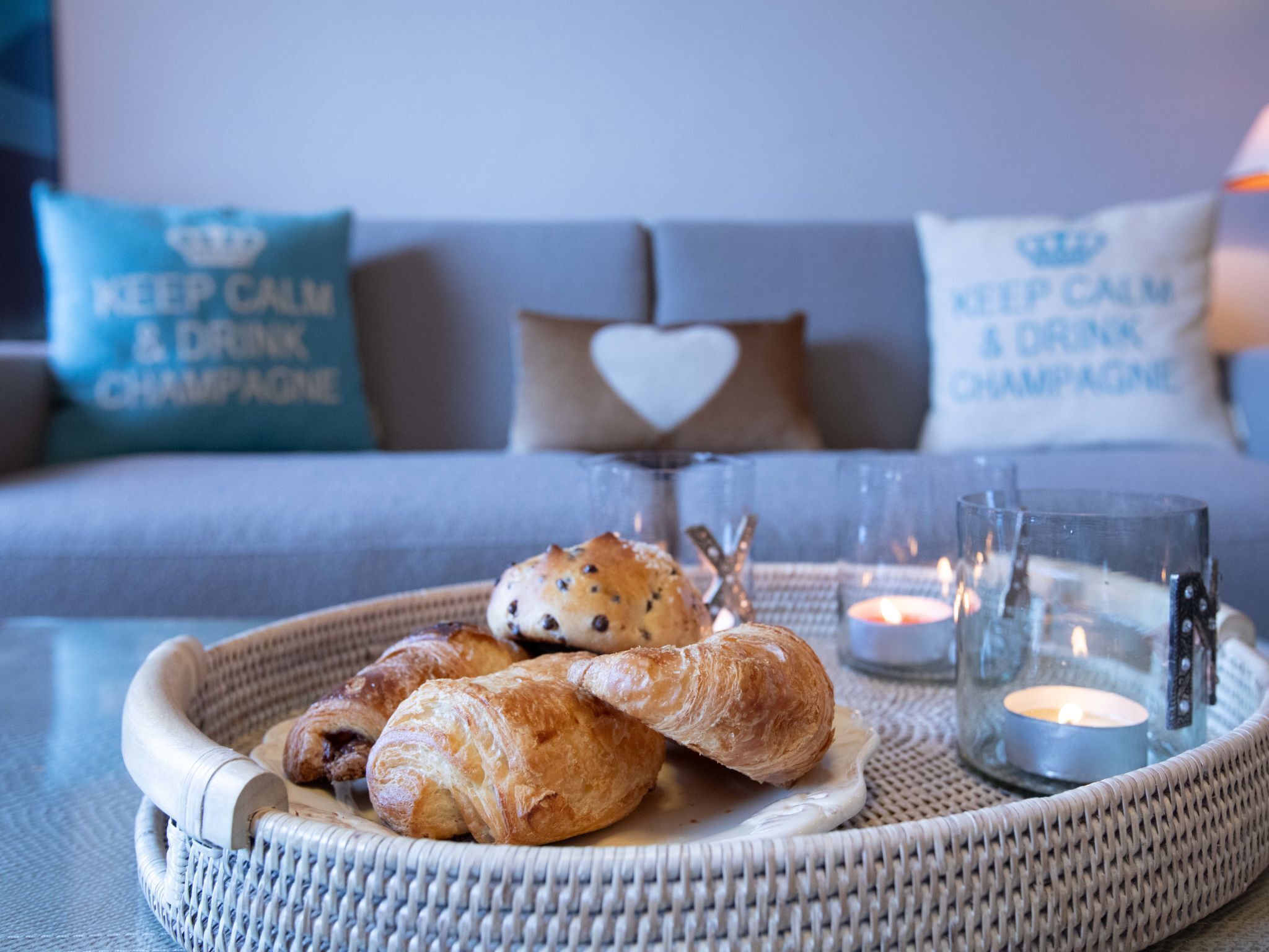 A wicker tray with croissants and a candle on a table, with a sofa and pillows in the background.