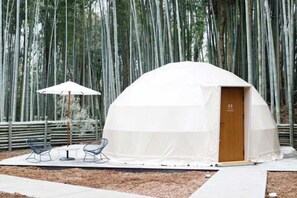 Una tienda de forma abovedada con una puerta, rodeada por un bosque de bambú. Hay dos sillas y una mesa con sombrilla.