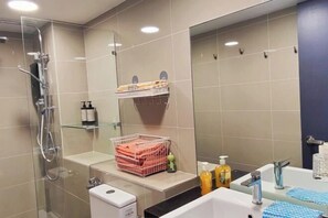 A modern bathroom with a glass shower enclosure, a sink with a mirror, and a shelf with towels and toiletries. - Chill Lah Homestay (Iskandar Puteri)