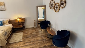 Een moderne slaapkamer met een houten vloer, een bed met hoofdeinde, twee blauwe stoelen, en een spiegel die een kamer met een bed en gordijnen weerspiegelt.