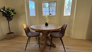 Ein runder Holztisch mit vier Stühlen, einer Topfpflanze und einer Blumenvase. Drei Fenster bieten Blick auf Bäume und einen klaren Himmel.
