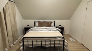 Ein kleines Zimmer mit einem Einzelbett, schrägen Decken, einem Fenster mit Vorhängen, einem Nachttisch mit Lampe und einem Beistelltisch mit Pflanze.