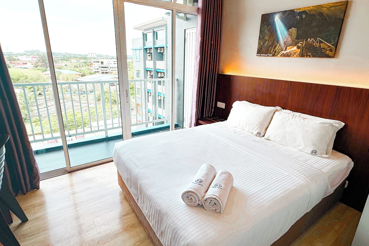 Habitación de hotel con una cama grande, un cuadro en la pared y un balcón con vistas a edificios y vegetación.