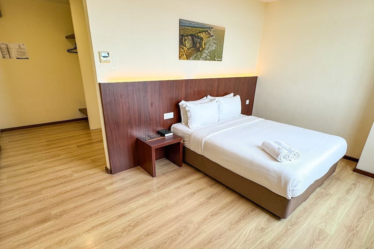 Una habitación de hotel con piso de madera, una cama con ropa blanca, una mesita de noche con un libro y una lámpara, y un cuadro en la pared.