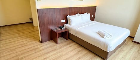 A hotel room with a wooden floor, a bed with white linens, a nightstand with a book and a lamp, and a painting on the wall.