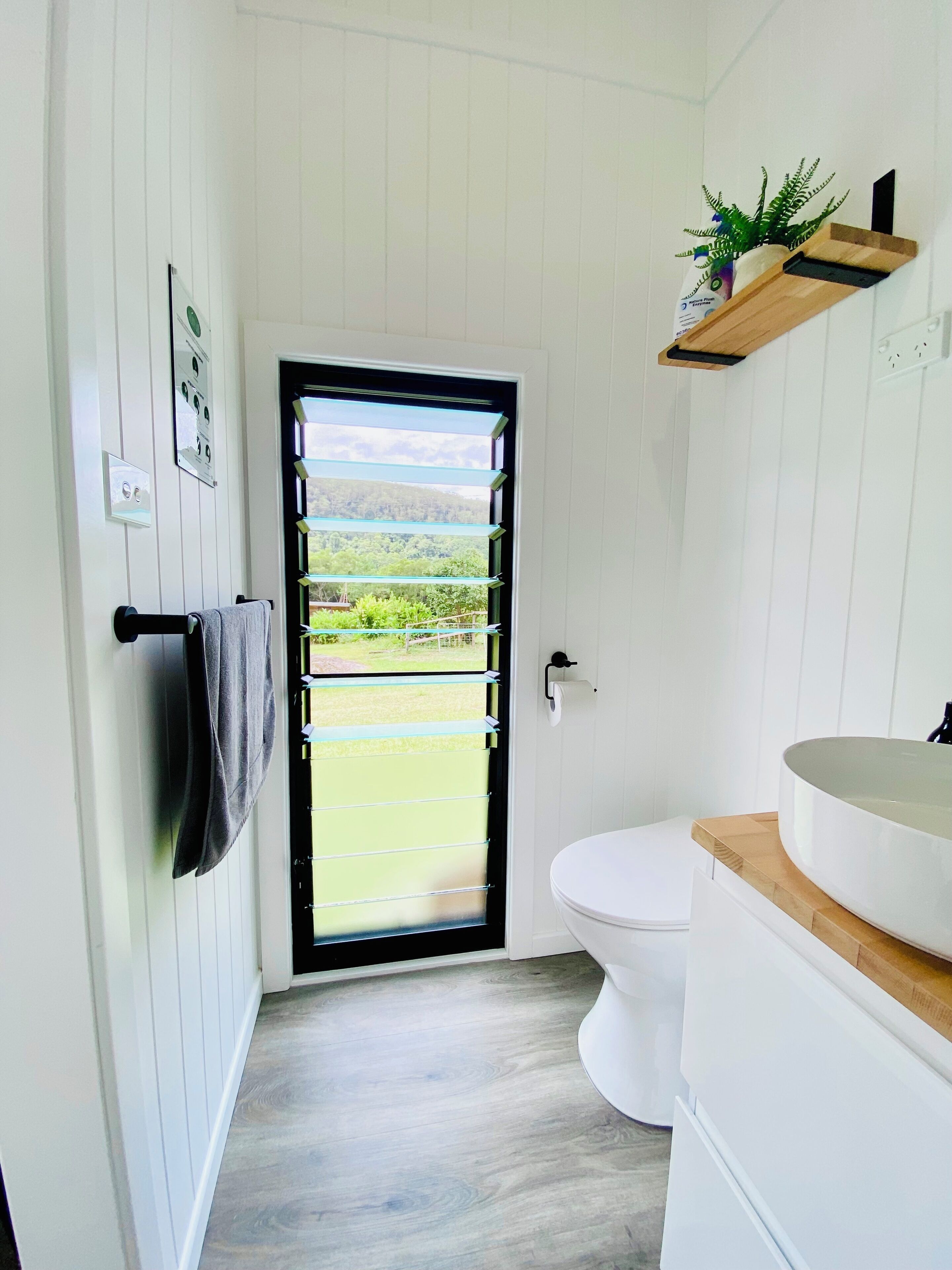Un baño con inodoro, lavamanos, toallero, ventana con vistas a follaje y estante con una planta.