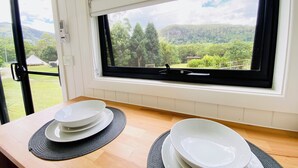 Une cuisine avec une fenêtre donnant sur un paysage verdoyant, un plan de travail en bois et un ensemble d’assiettes blanches disposées sur un set de table.