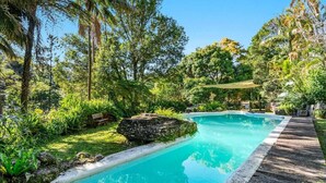 Une piscine rectangulaire entourée d’une terrasse en bois, située dans un jardin luxuriant agrémenté de divers arbres et plantes.