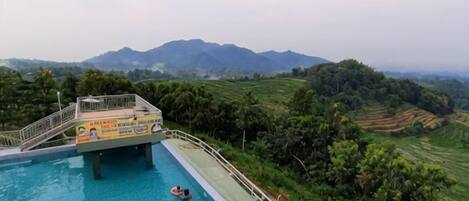 A swimming pool with a bridge, surrounded by greenery and mountains.