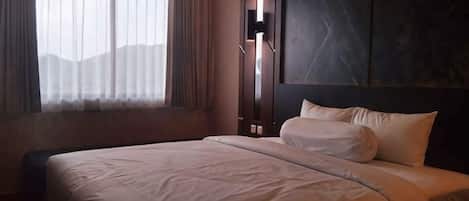 A hotel room with a large bed, a bedside table, a lamp, and a view of the outdoors through the window.