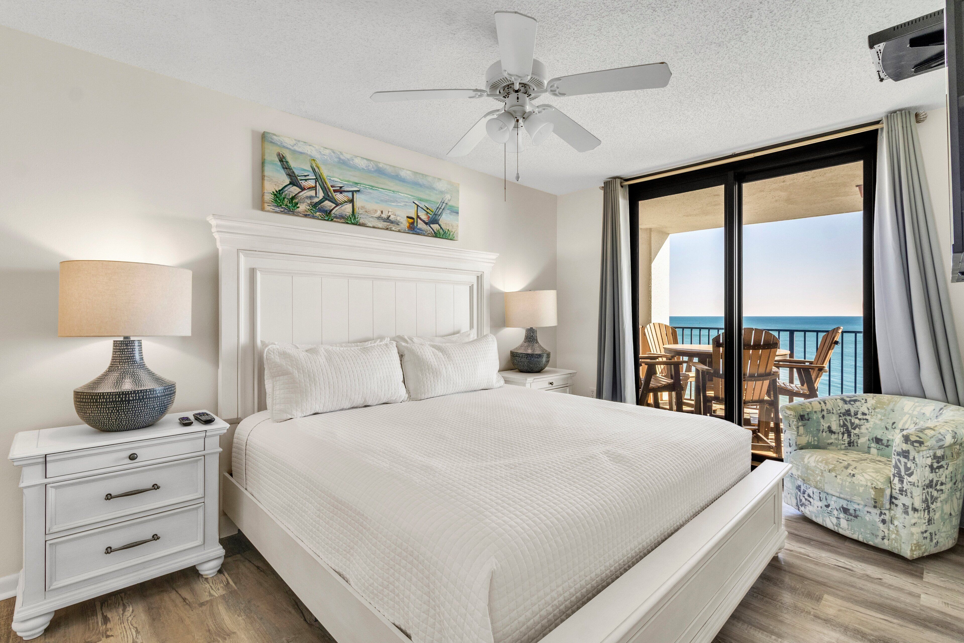 A hotel room with a large bed, white nightstands, a ceiling fan, a painting, and a balcony with chairs.