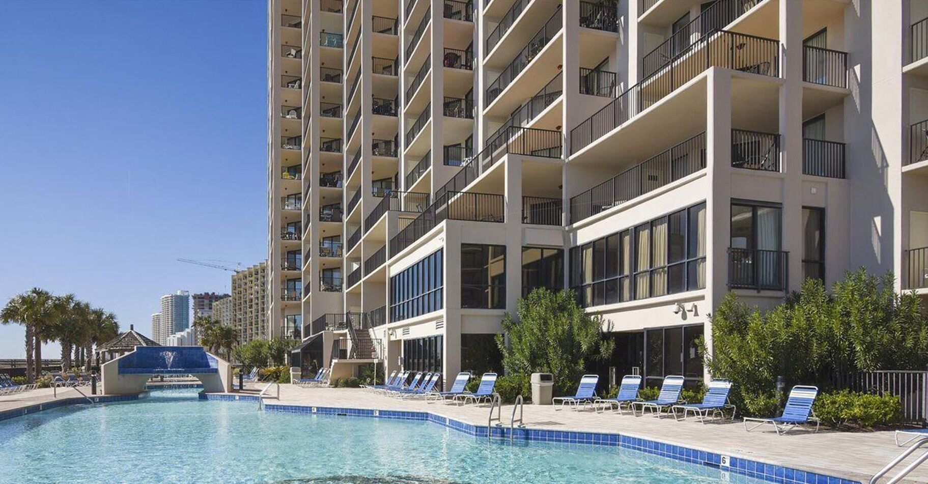 A hotel with a swimming pool, multiple balconies, and a clear sky.