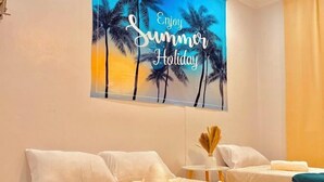 Quarto de hotel com cama, almofadas, uma mesinha com um vaso e decoração na parede com palmeiras e o texto 'Enjoy Summer Holiday'.