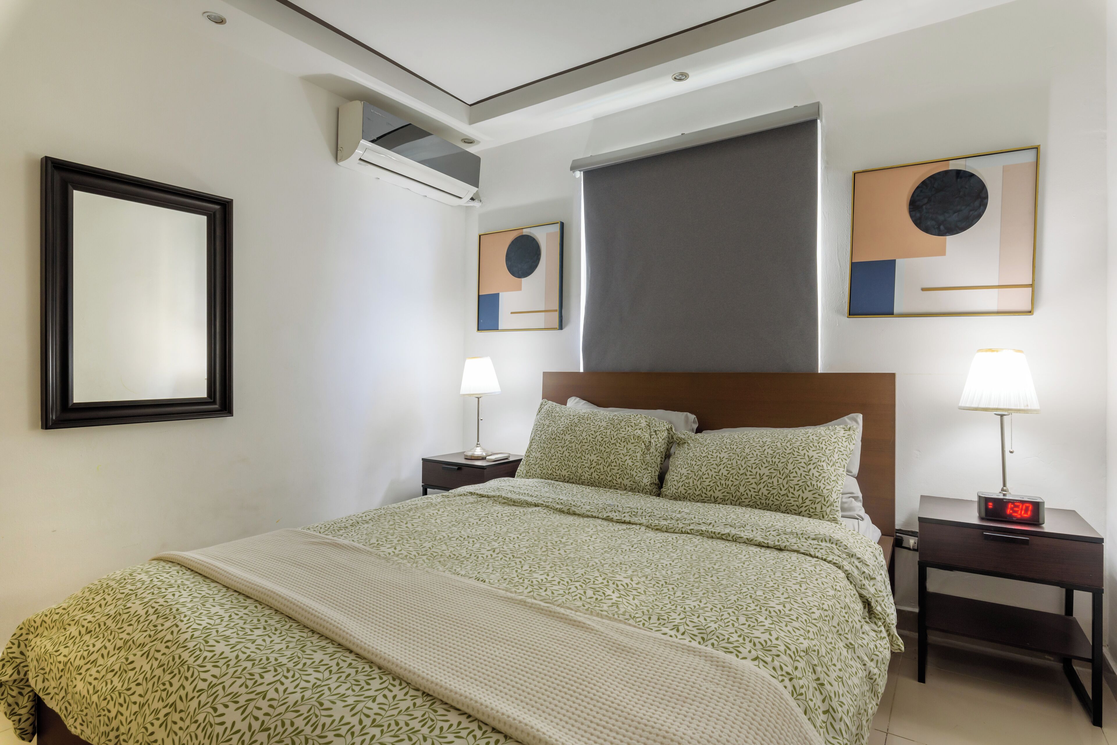 A hotel room with a bed, two bedside tables with lamps, a large window with a blind, and a wall-mounted air conditioner.