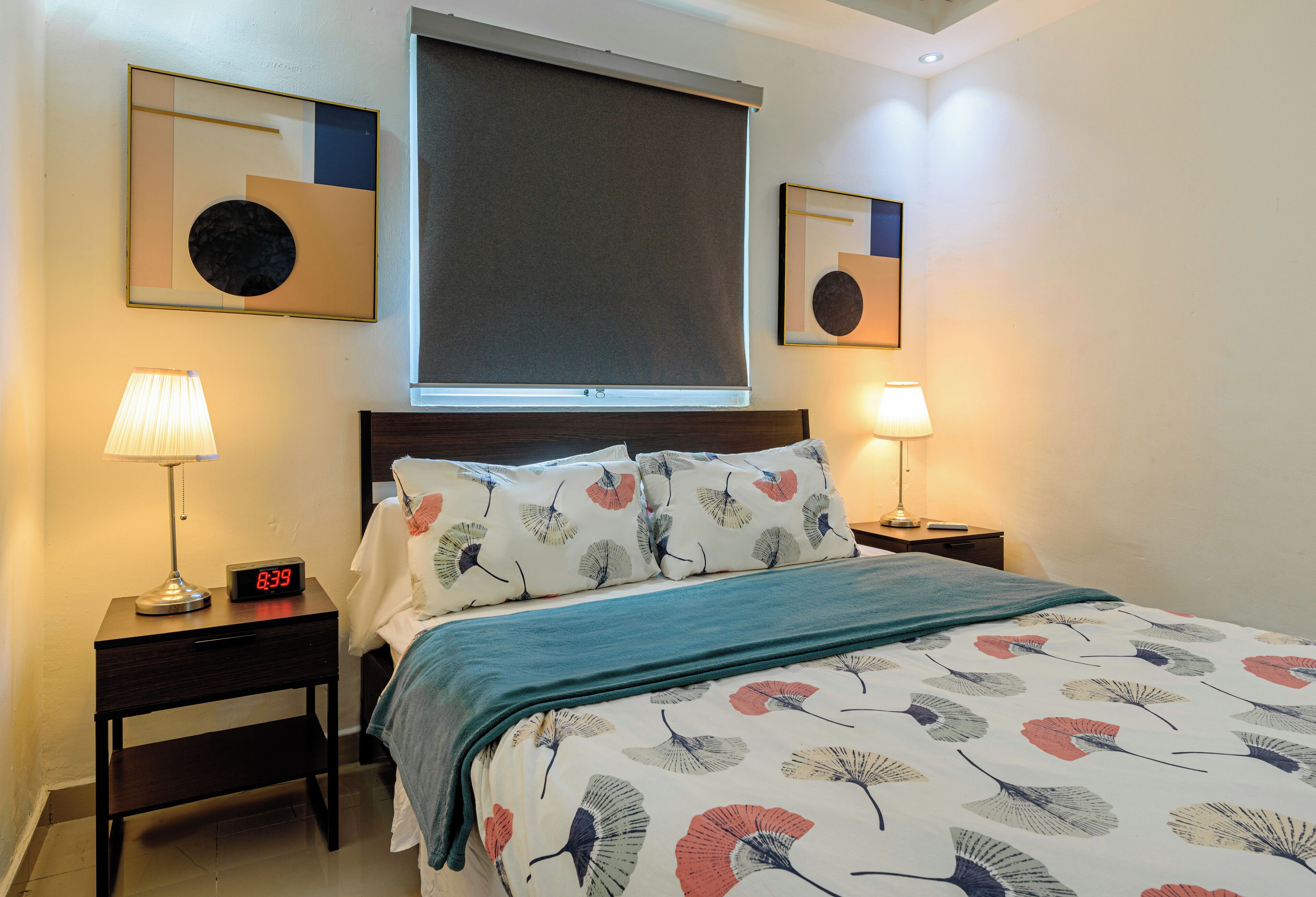 Cama bem arrumada com colcha florida, duas luminárias de cabeceira, um relógio digital e duas obras de arte emolduradas na parede.