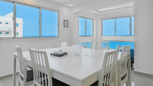Un espace repas moderne avec une grande table et des chaises blanches, de grandes fenêtres offrant une vue sur la mer et un tableau encadré au mur.