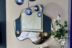 Quarto com espelho de design único, cama com roupa de cama branca e planta decorativa com flores brancas.