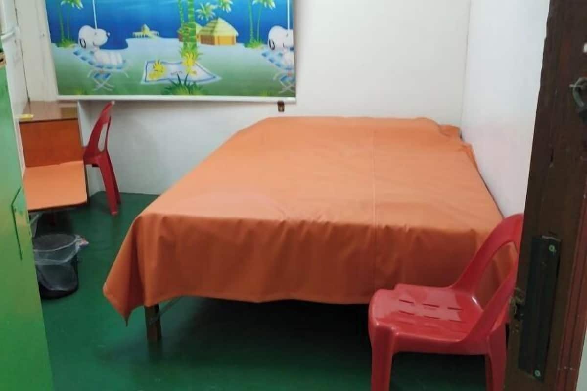 Ένα μικρό δωμάτιο με ένα μονό κρεβάτι, μια κόκκινη πλαστική καρέκλα, ένα μικρό τραπέζι και έναν πίνακα κρεμασμένο στον τοίχο.