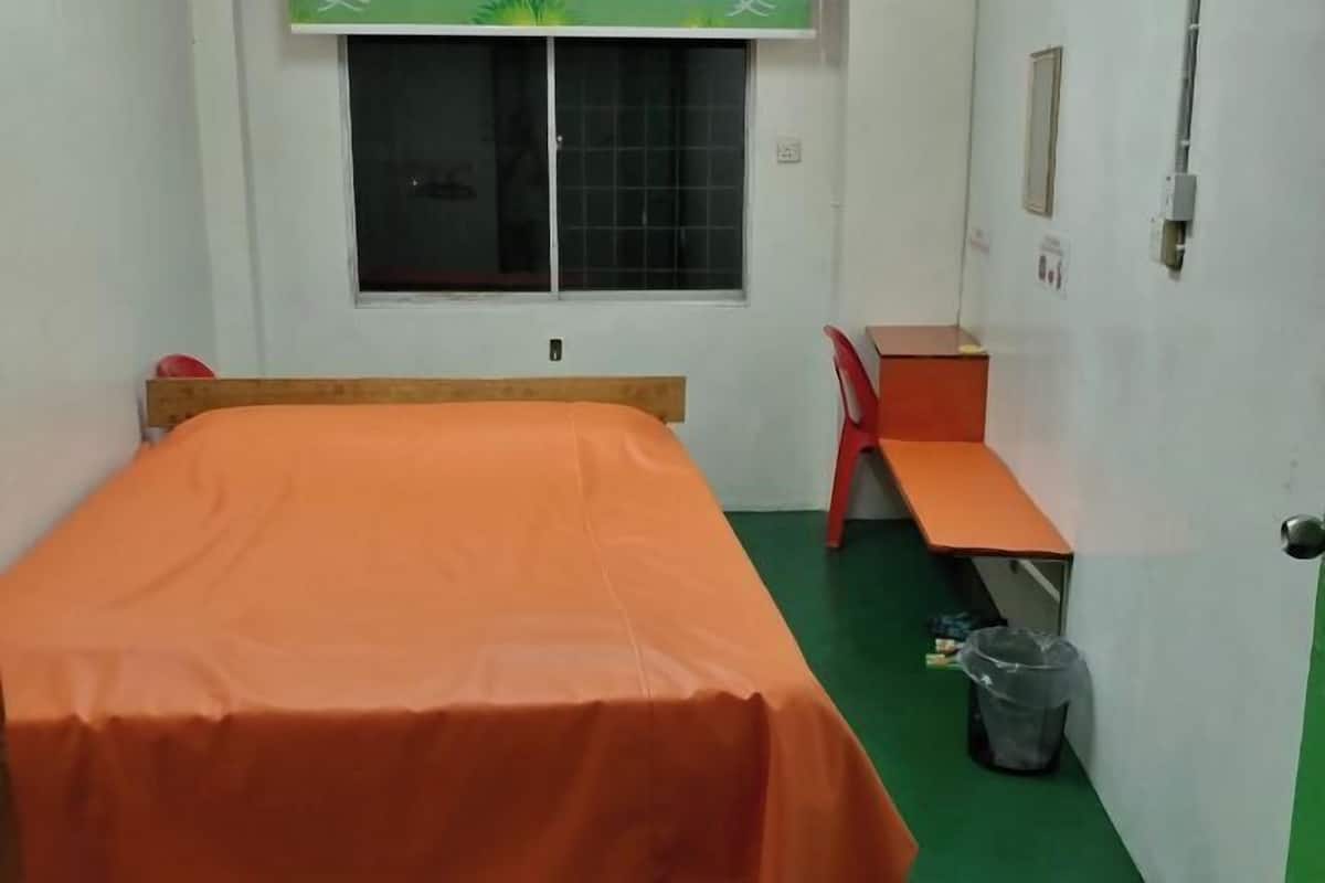 Ένα δωμάτιο με ένα μονό κρεβάτι, ένα πορτοκαλί κάλυμμα κρεβατιού, ένα παράθυρο με σχέδιο σε σχήμα πλέγματος, ένα γραφείο με καρέκλα και έναν κάδο απορριμμάτων.