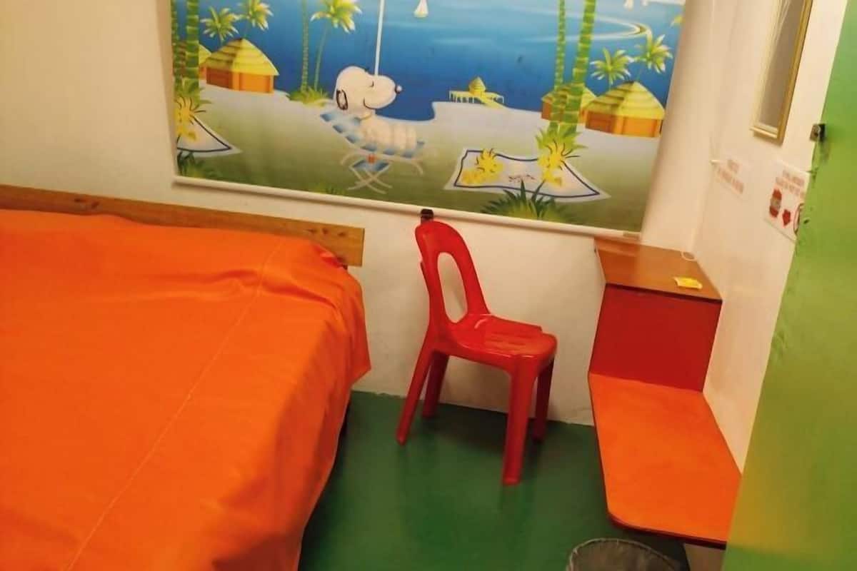 Ένα δωμάτιο με δύο κρεβάτια, μια κόκκινη πλαστική καρέκλα, ένα μικρό τραπέζι και μια τοιχογραφία.