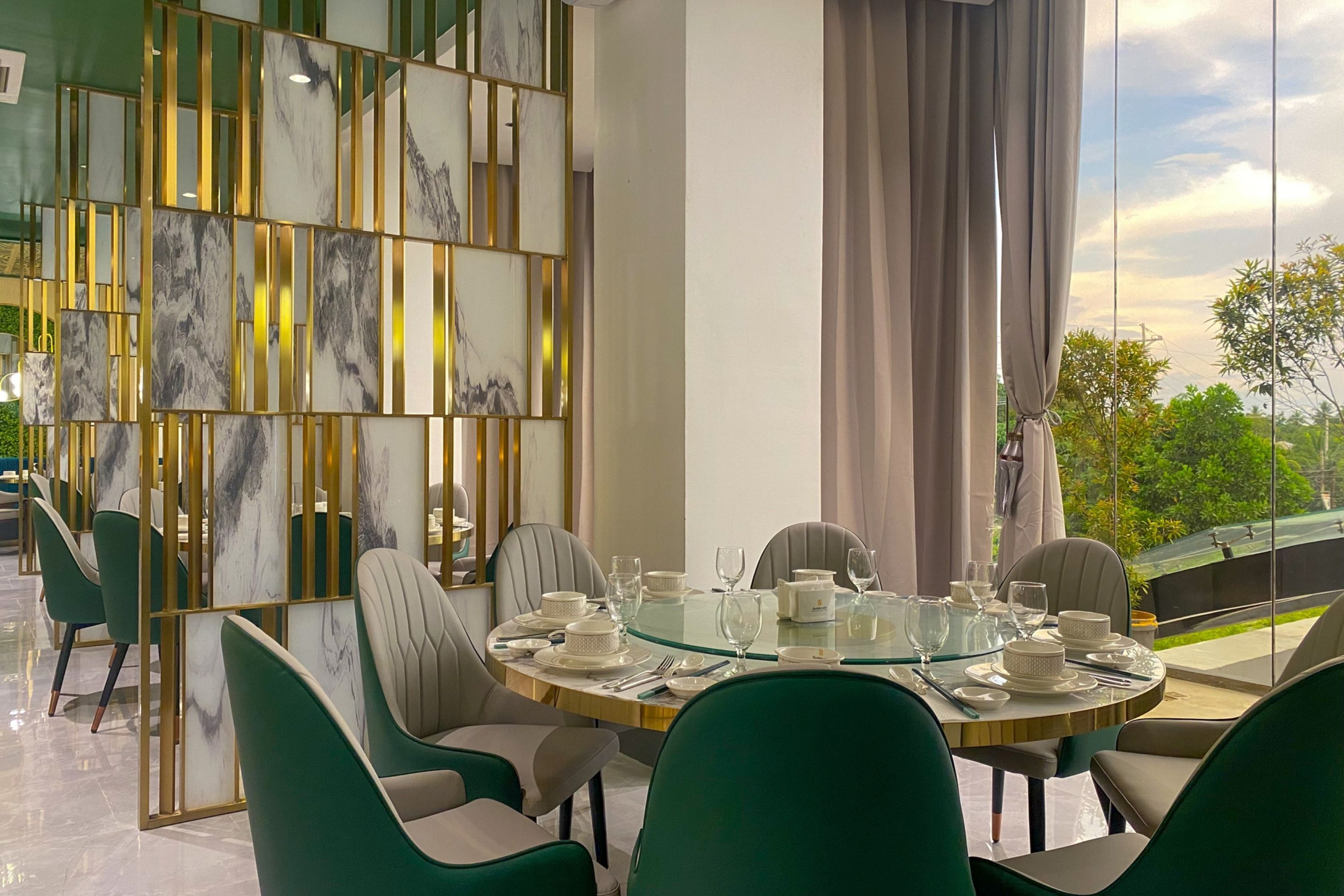 A dining area with a round glass table, green upholstered chairs, and a decorative wall with vertical gold elements and marble panels.