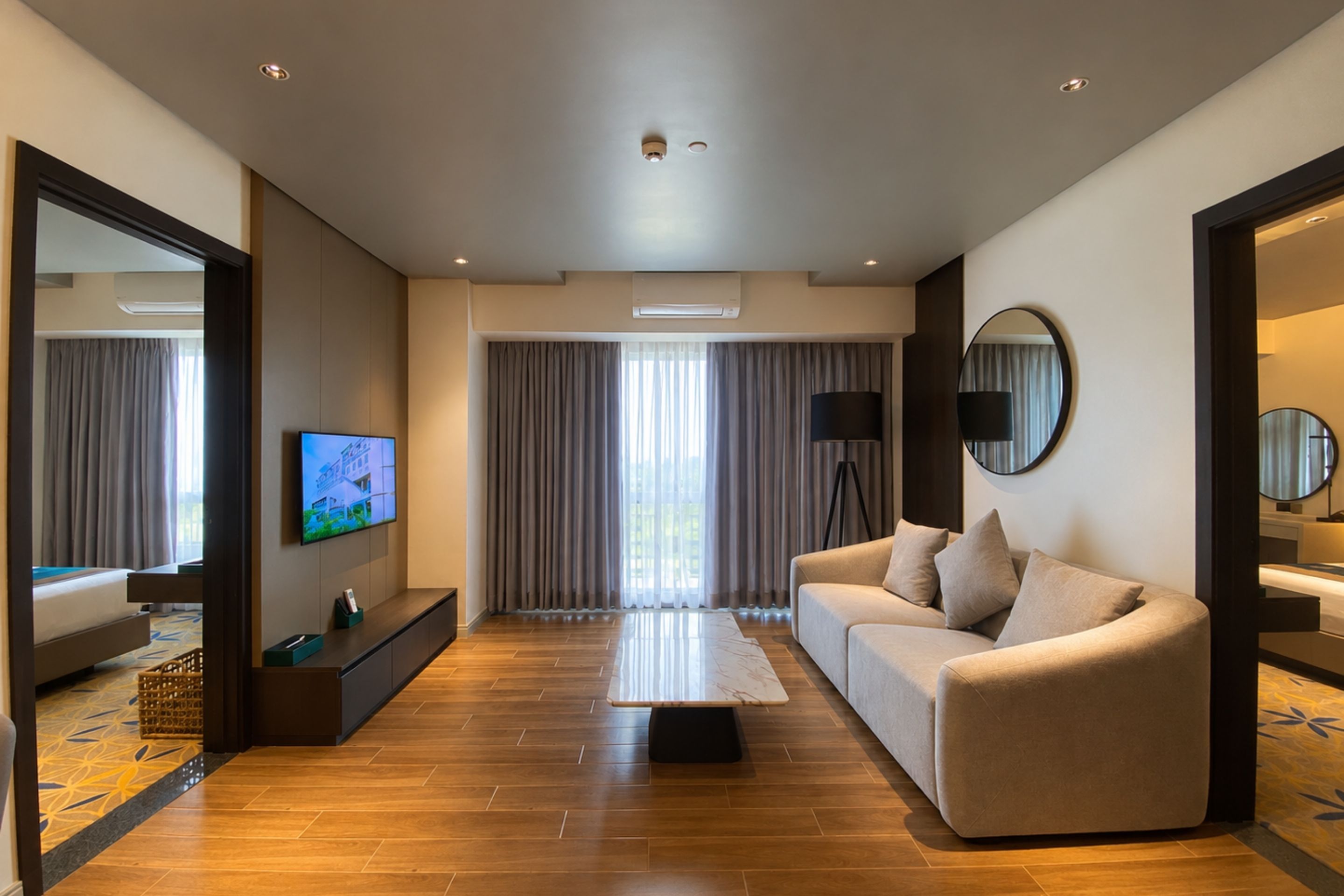 A modern hotel room with a sofa, a TV, a coffee table, and a large window with curtains.