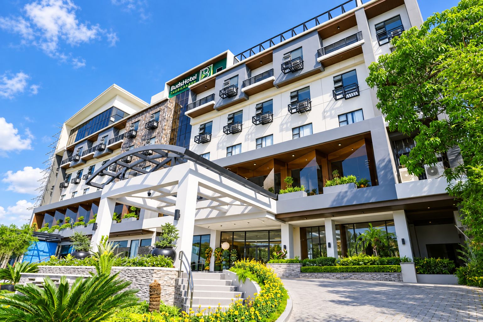 A multi-story hotel with a modern design, featuring balconies with plants, a covered walkway, and a clear sky.