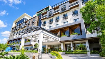 A multi-story hotel with a modern design, featuring balconies with plants, a covered walkway, and a clear sky.