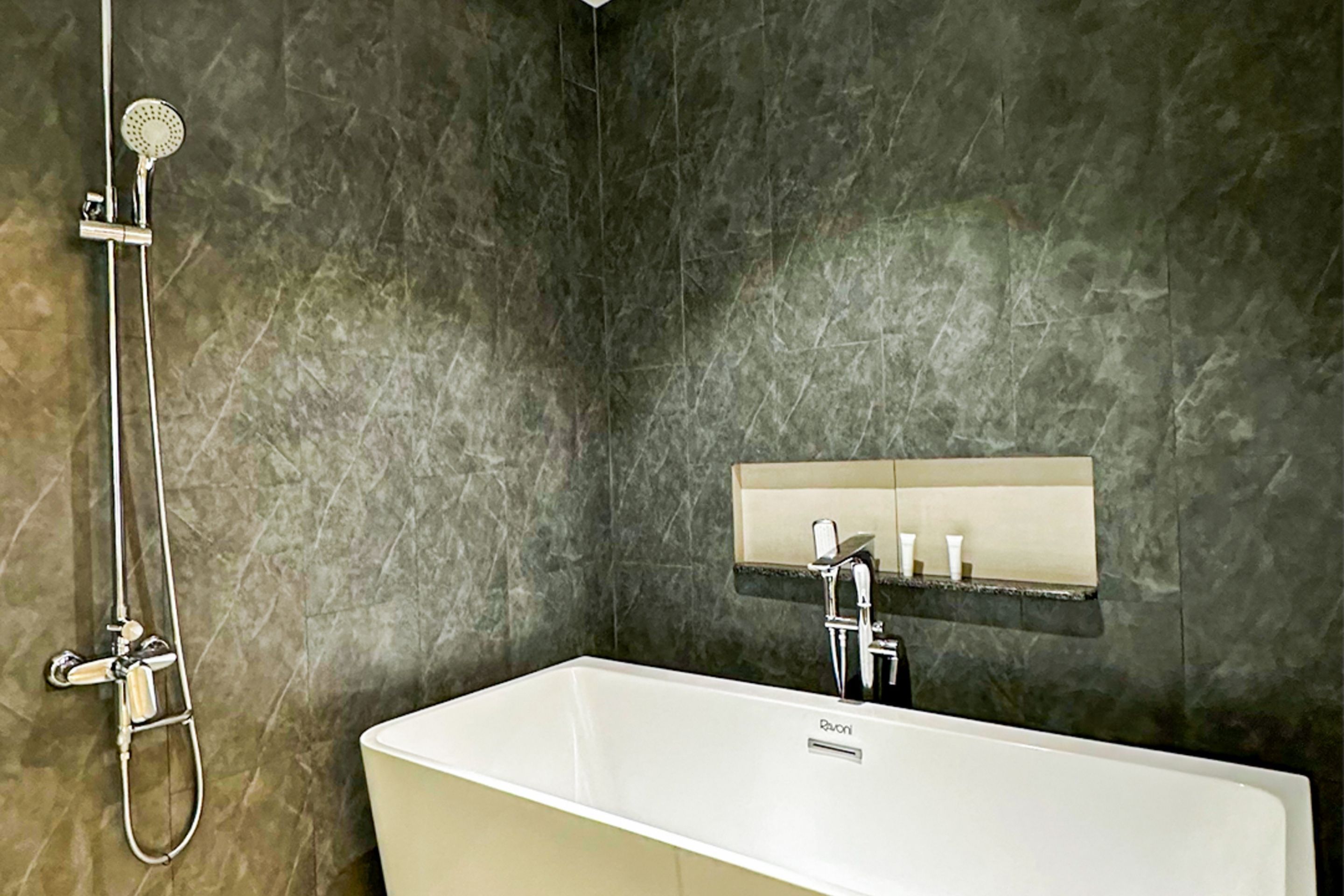 Een badkamer met een wit bad, een handdouche en een spiegel aan de muur met een plank erboven.