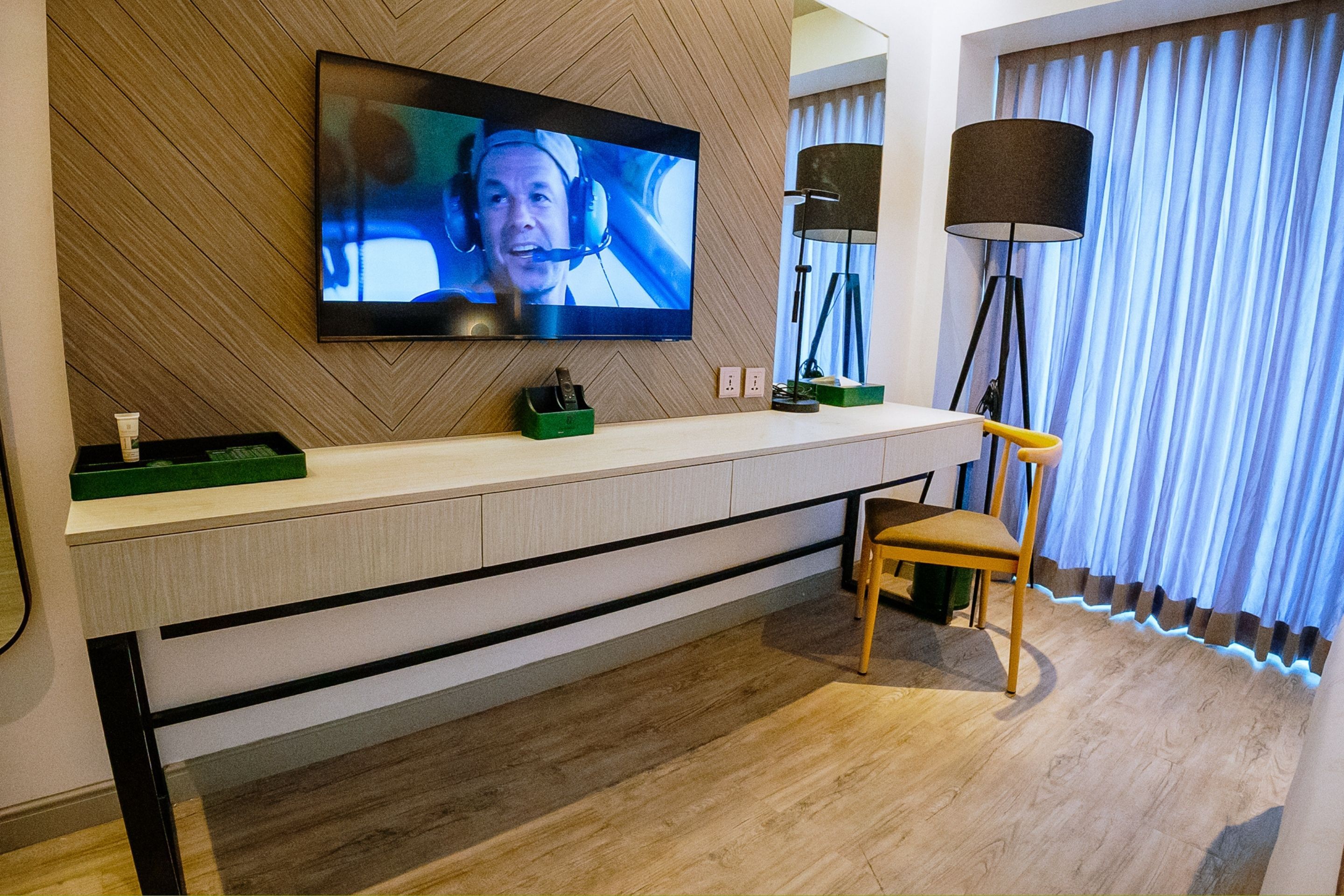 A modern hotel room with a flat-screen TV mounted on a wooden wall, a wooden chair, a bedside table with a lamp, and a large window with curtains.