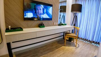 A modern hotel room with a flat-screen TV mounted on a wooden wall, a wooden chair, a bedside table with a lamp, and a large window with curtains.
