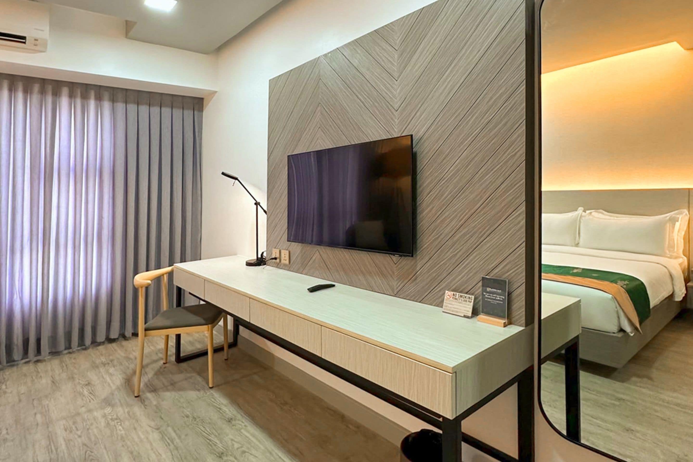 A hotel room with a desk, chair, television, and a bed with a green throw.