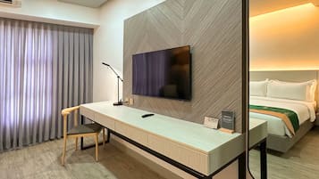 A hotel room with a desk, chair, television, and a bed with a green throw.