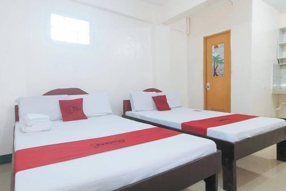 Habitación de hotel con dos camas individuales, cada una con una colcha rojo y blanco, almohadas blancas y una almohada roja. Se observa una puerta de madera con una pintura de palmera, un ventanal pequeño y una pared blanca.