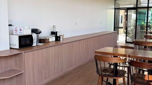 A modern kitchen area with a microwave, coffee maker, and other appliances on a counter. There are wooden tables and chairs arranged for dining. - The Model Garden (Yasothon)