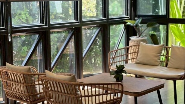 A room with large windows, wicker furniture, a wooden table, and a cushioned sofa. - The Model Garden (Yasothon)